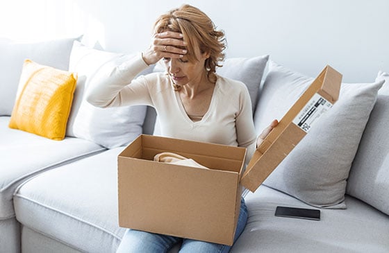 Person sitting on a couch looking at an unexpected online purchase delivery, illustrating eCommerce uniqueness scams and online shopping fraud.